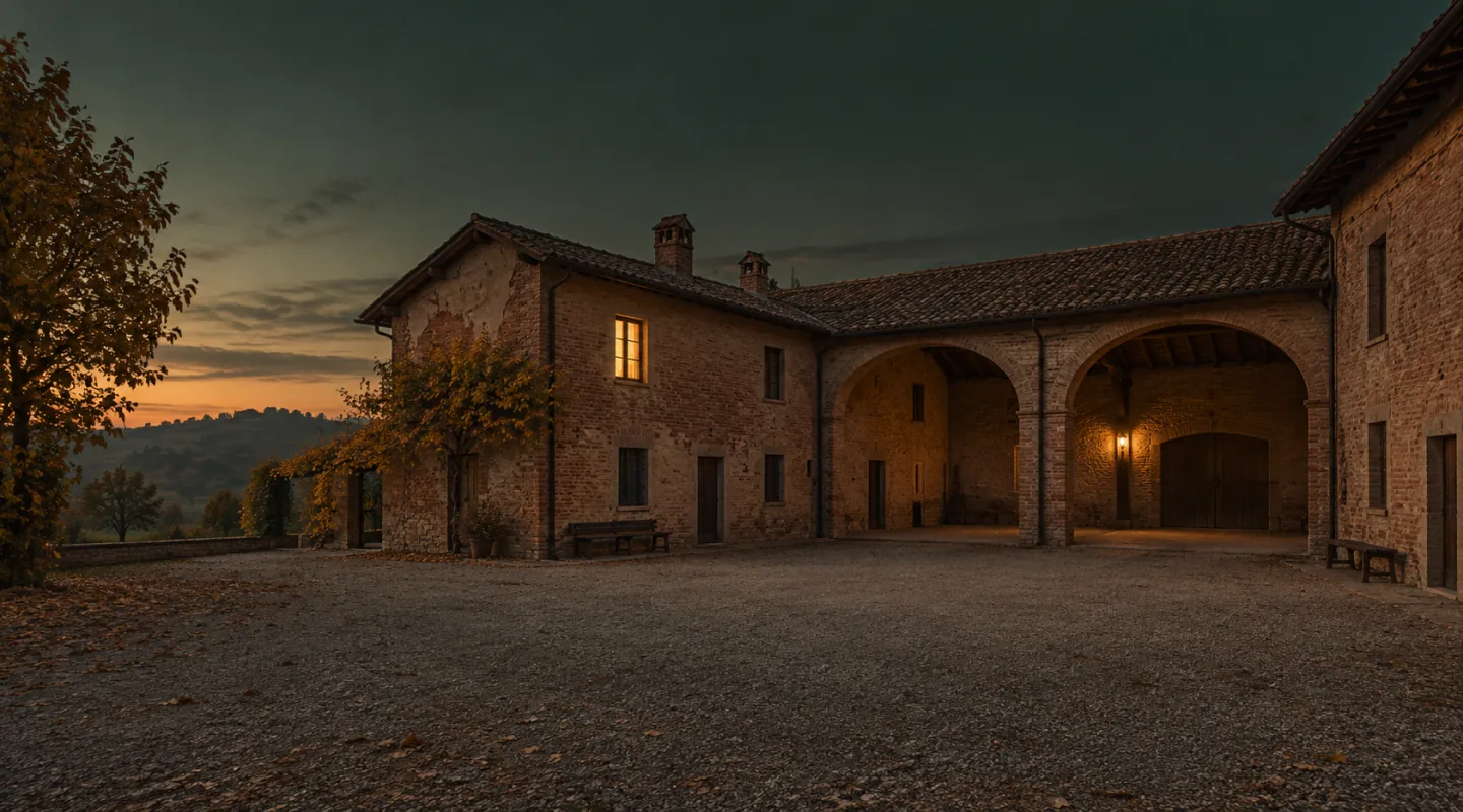 Cortile di una cascina deserto all’imbrunire: muri di mattoni, un portico ad archi, una panca di legno vuota e la luce calda di una finestra